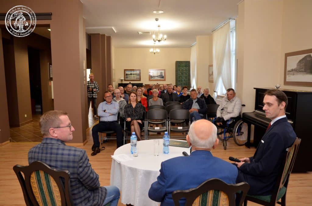 Otwarcie wystawy "Pamiętne eliminacje" w Muzeum Solca. Na pierwszym planie, siedzący przy stole i odwróceni tyłem pracownicy Muzeum wraz z gościem Andrzejem Strejlauem. Dalej siedząca publiczność.