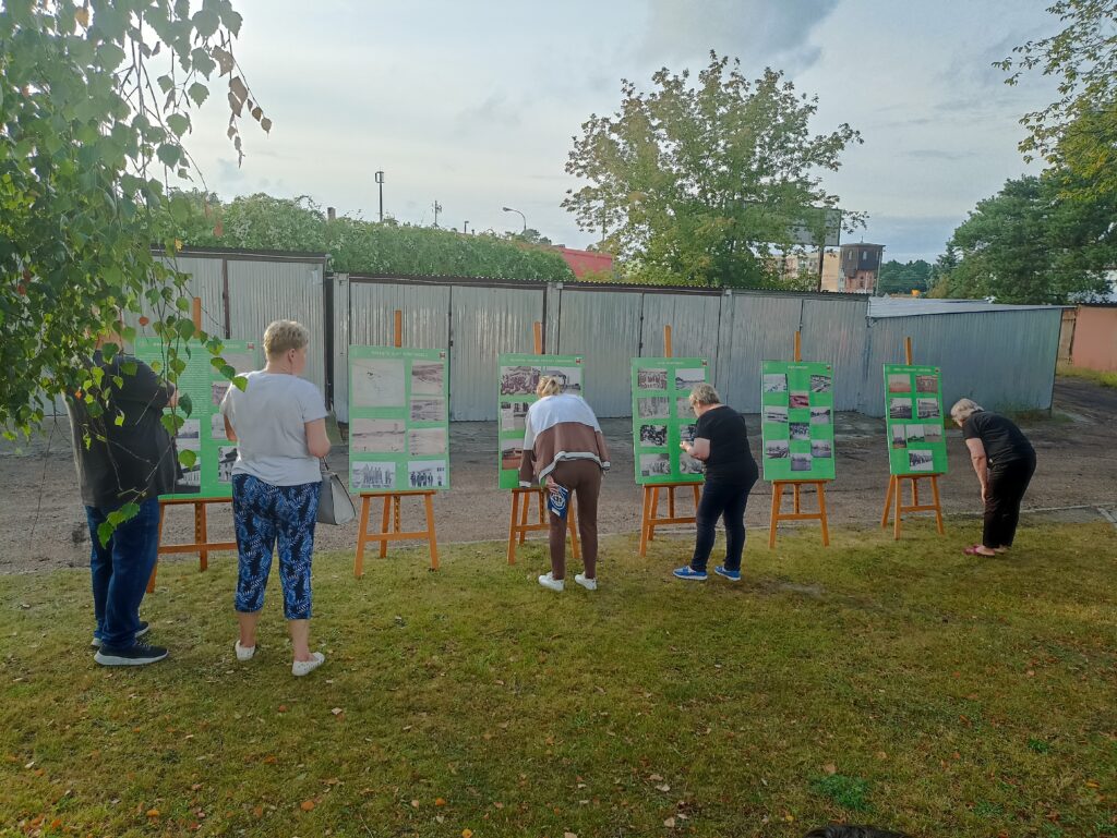 Pięć osób odwróconych tyłem do aparatu, ogląda zielone plansze informacyjne z wystawą plenerową, podczas pikniku rodzinnego osiedla robotniczego.
