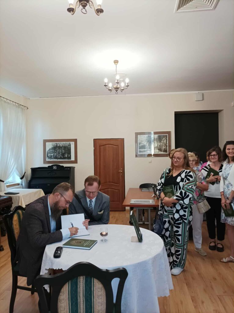 Pan Marcin Hlebionek siedzi przy okrągłym stole i podpisuje książkę. Po prawej stronie zdjęcia kolejka osób chcących zdobyć autograf autora monografii.