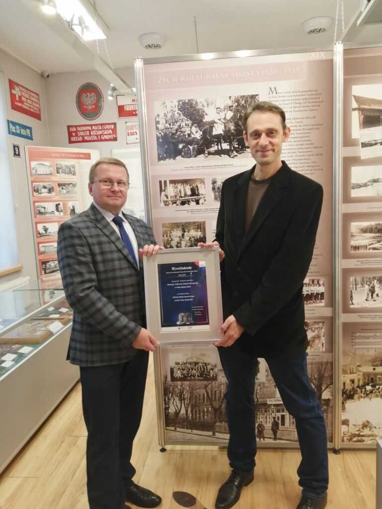 Dyrektor Muzeum Rafał Kubiak oraz Kustosz Łukasz Wojtecki pozujący z wyróżnieniem w konkursie MUSEJON – Kujawsko-Pomorskie Muzealne Wydarzenie Roku, na tle sali wystawowej Muzeum Solca.