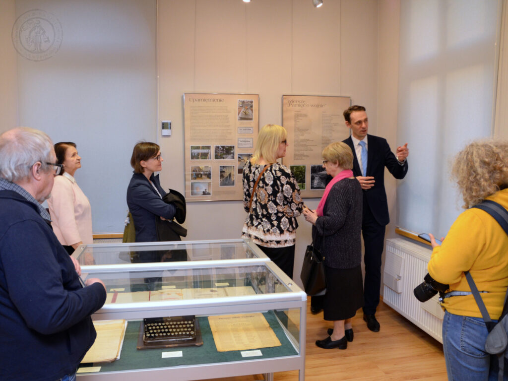 Zdjęcie przedstawia małą salę wystaw czasowych, gdzie Łukasz Wojtecki przybliża grupie osób historię pisarki.